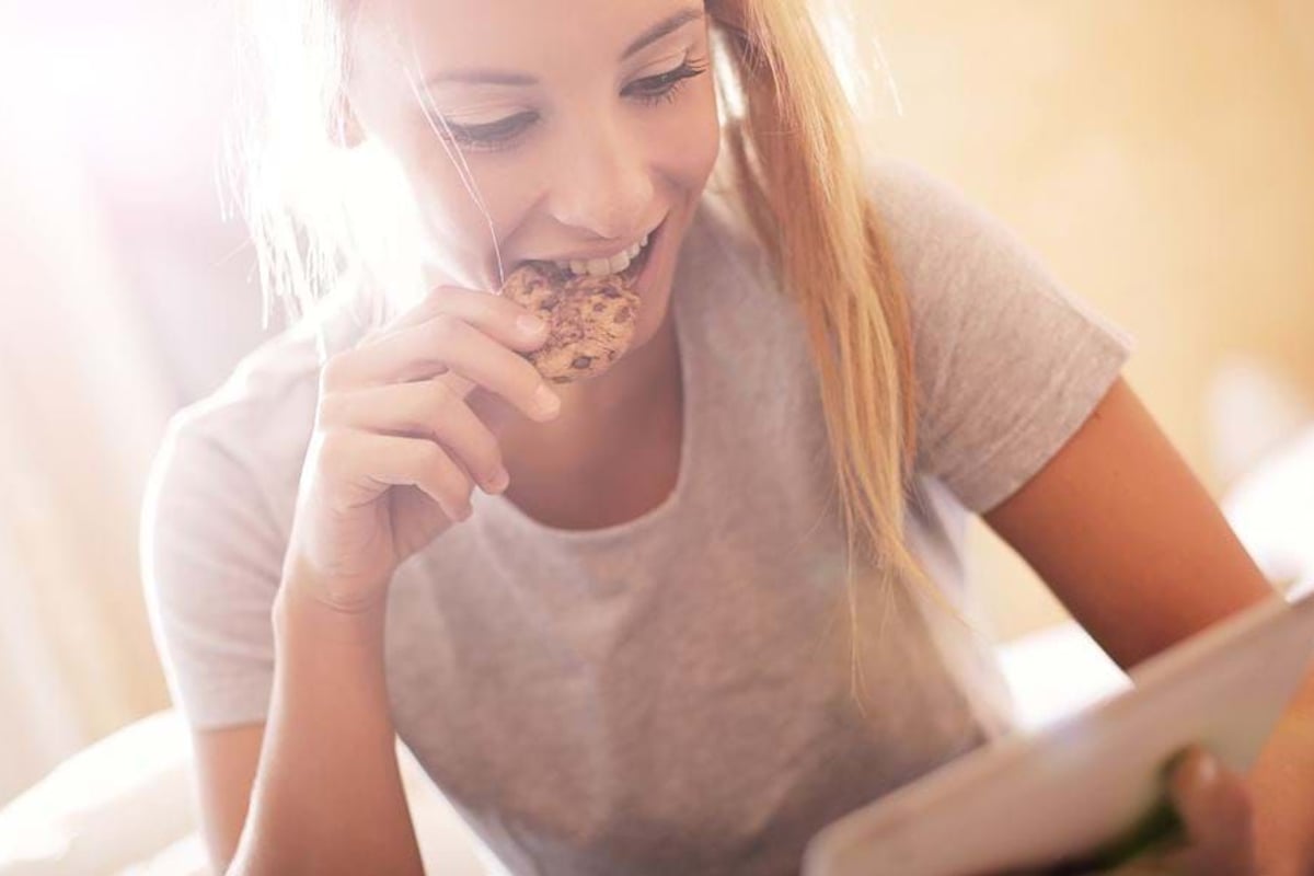 Young woman eating a cookie while looking at a tablet in bright sunlight