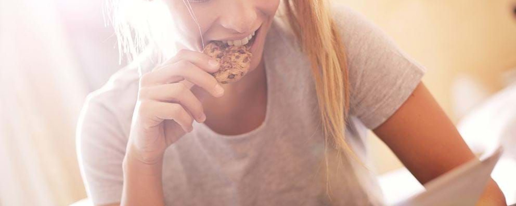 Young woman eating a cookie while looking at a tablet in bright sunlight