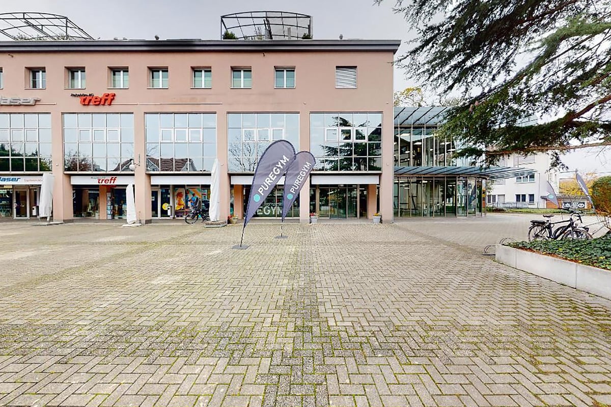 PureGym Lyss: Place extérieure avec porte-vélos, zone pavée et bâtiment à plusieurs étages avec façade commerciale.
