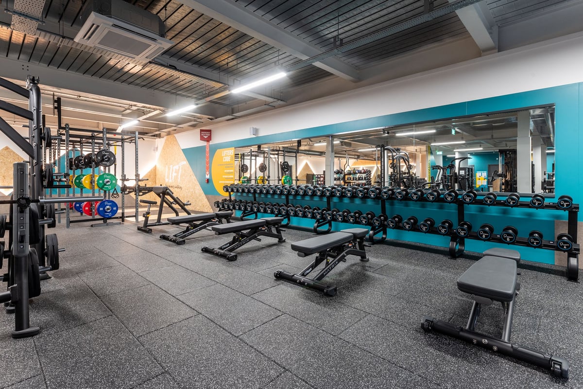 PureGym Droitwich Spa: Rows of weight benches, dumbbells, squat racks, and teal accent walls in a spacious gym facility.