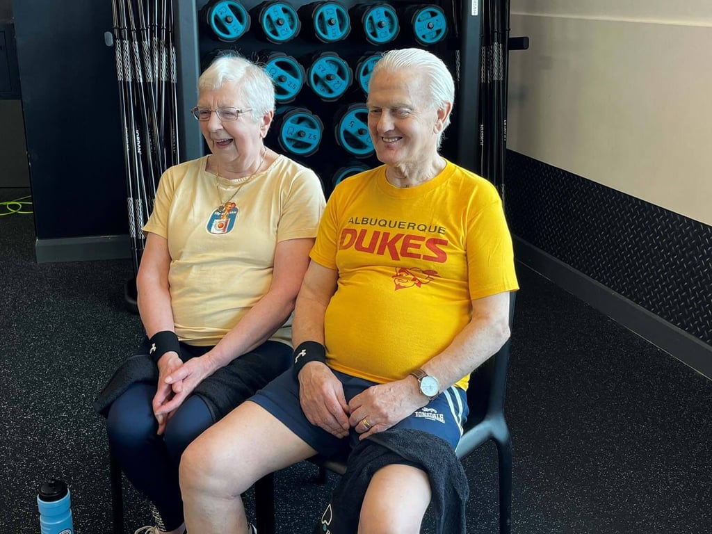 Two elderly people sitting on chairs in a gym with weights behind them smiling
