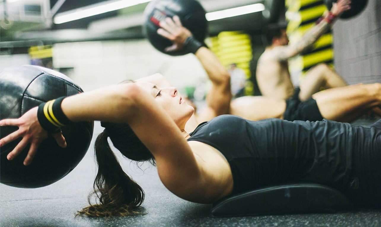 Woman exercising with a medicine ball lying on a mat in a gym with others in the backgroun