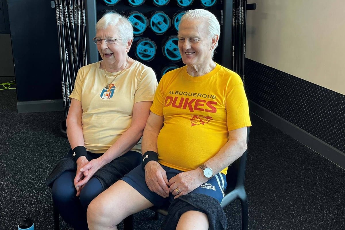 Two elderly people sitting on chairs in a gym with weights behind them smiling