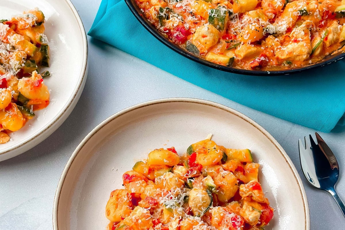 Two plates and a pan of gnocchi with vegetables and grated cheese on a blue napkin