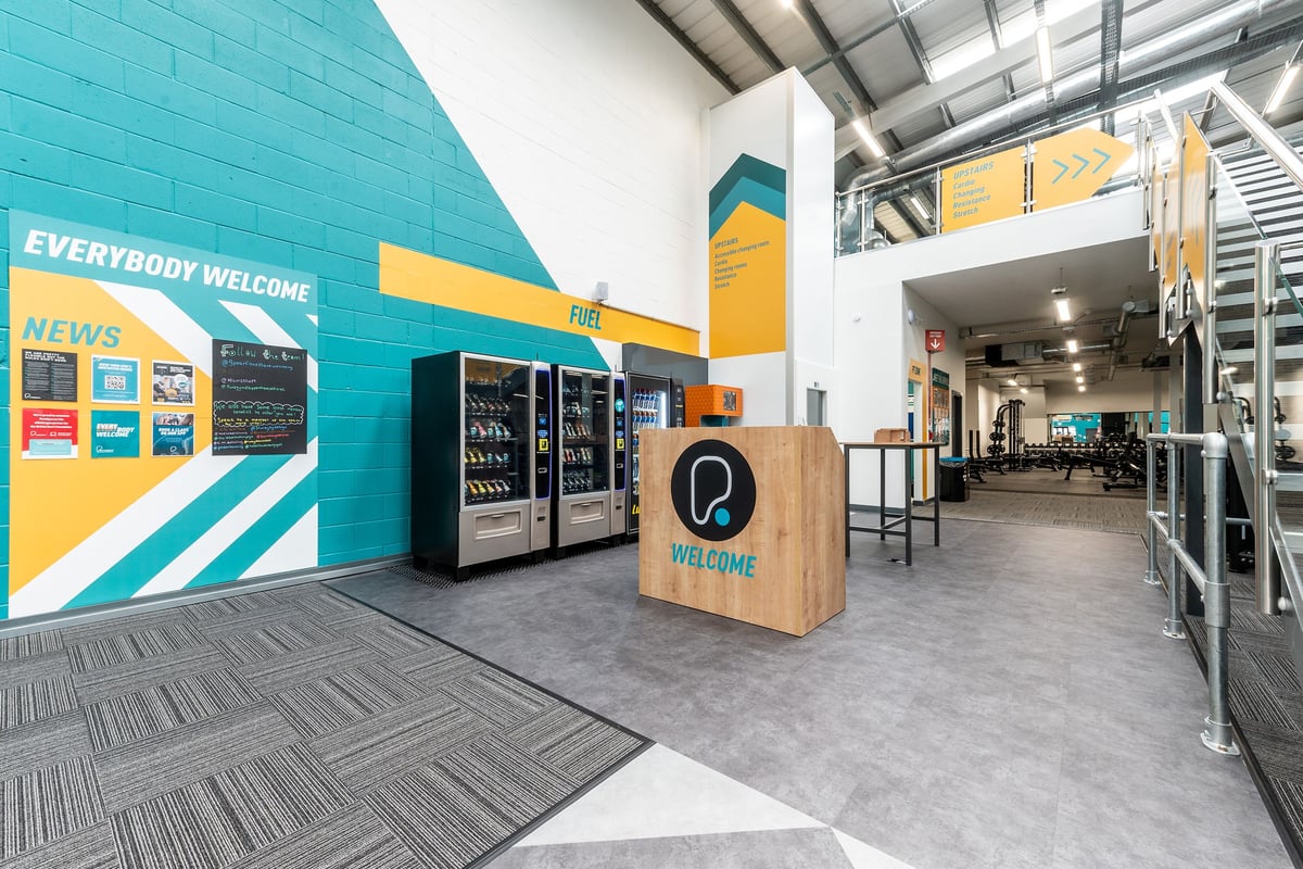 PureGym Chippenham: Vending machines, self-service kiosks, tables, and turquoise tiled walls inside a fitness center lobby.