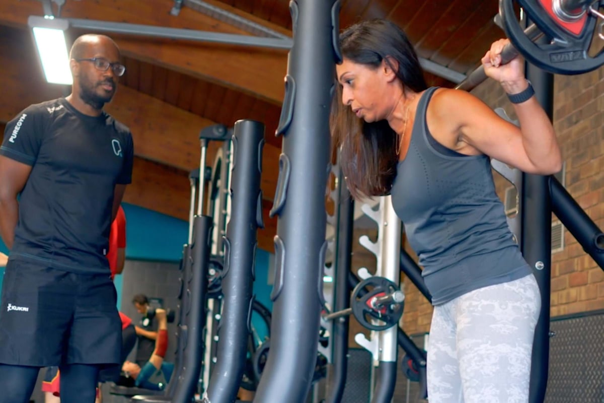 Woman lifting barbell on shoulders with trainer watching in gym setting
