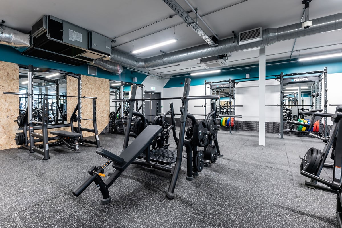PureGym Doncaster: Squat racks, bench presses, free weights, and rubberized flooring in a workout studio with ducts overhead.