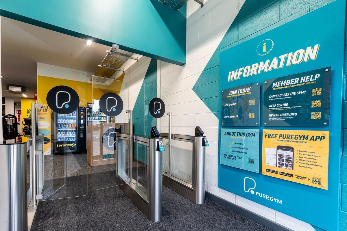 PureGym Dungannon: Entrance area with self-service kiosks, turnstiles, and information screens advertising gym app.