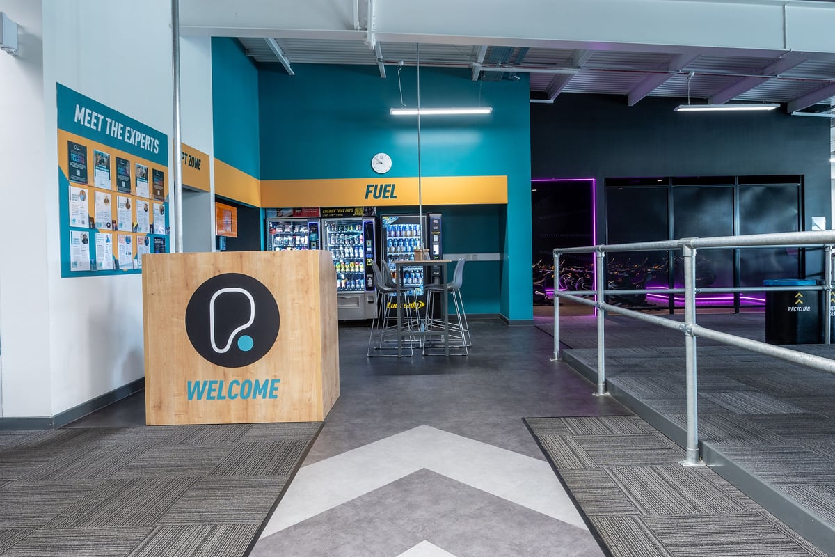 PureGym Manchester Cheetham Hill Retail Park: Cafe style seating, vending machines, and a welcome sign in a modern space.
