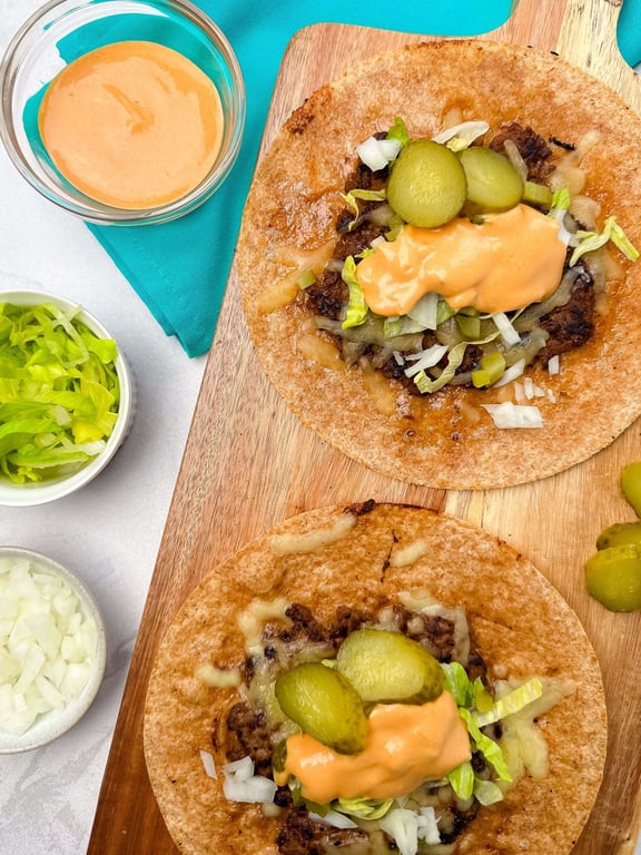 Two open wraps with beef, cheese, pickles, lettuce, onions, and sauce on a wooden board.