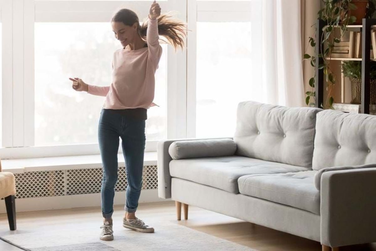 Woman dancing joyfully in a bright living room with a grey sofa and plants.