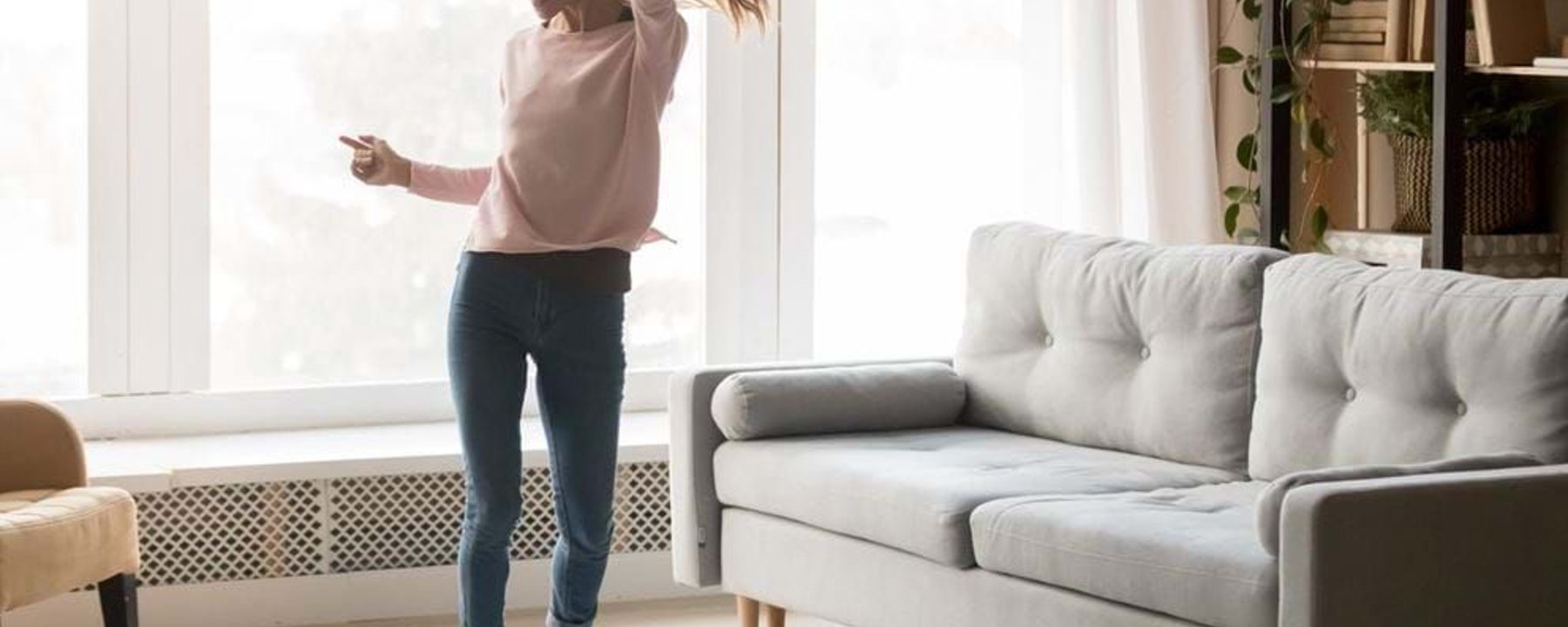 Woman dancing joyfully in a bright living room with a grey sofa and plants.