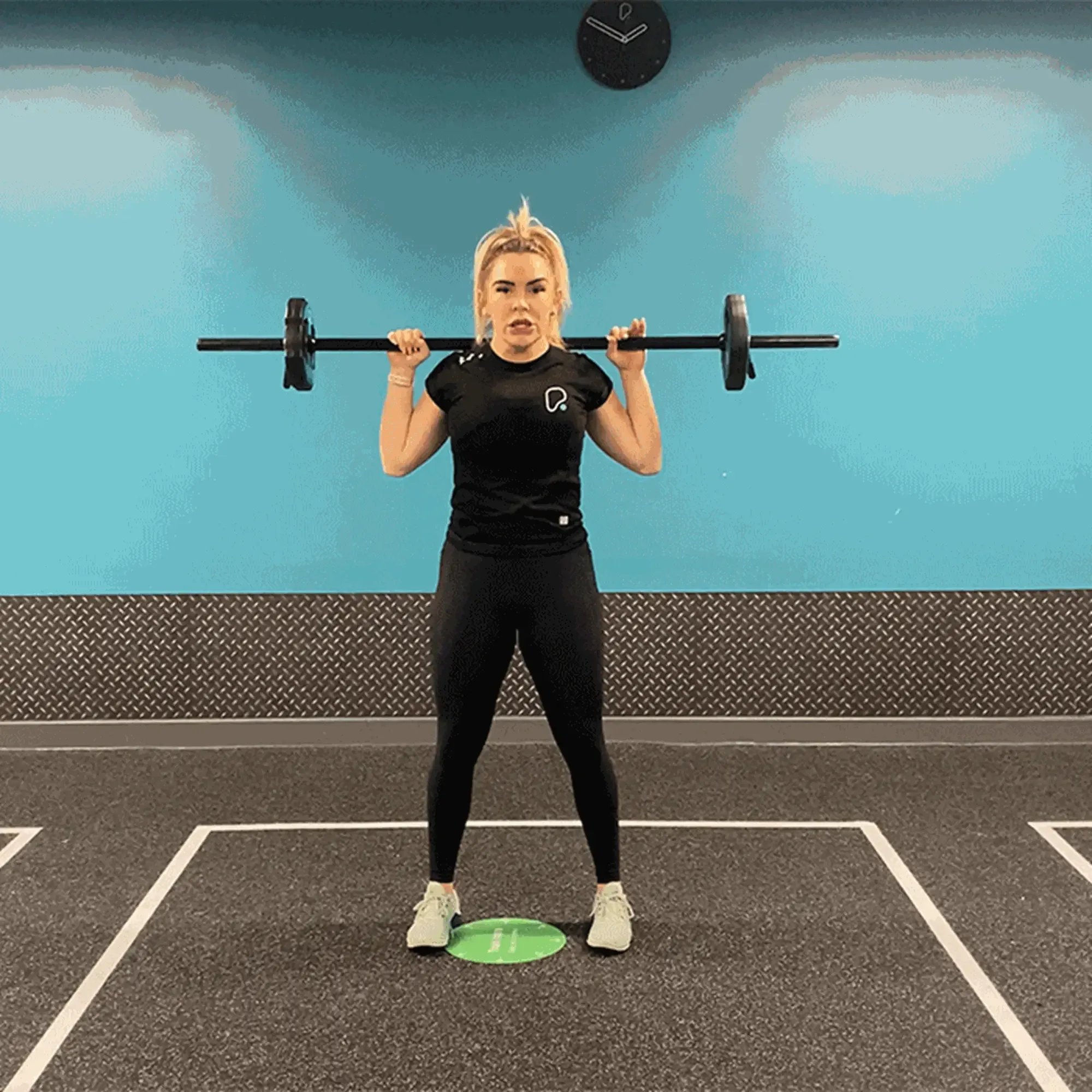 Woman in black sportswear lifting a barbell in a gym with blue walls and clock above