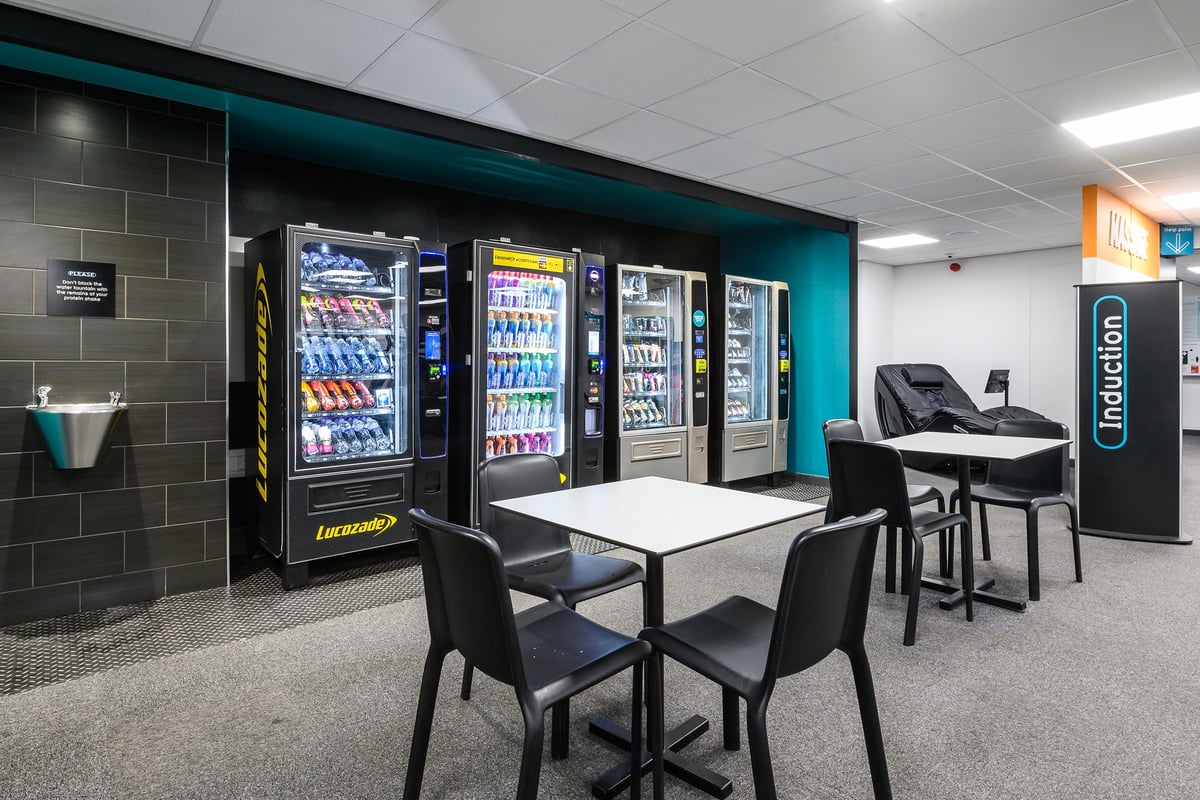 PureGym Burnham: Vending machines, tables and chairs in a room with teal accents.