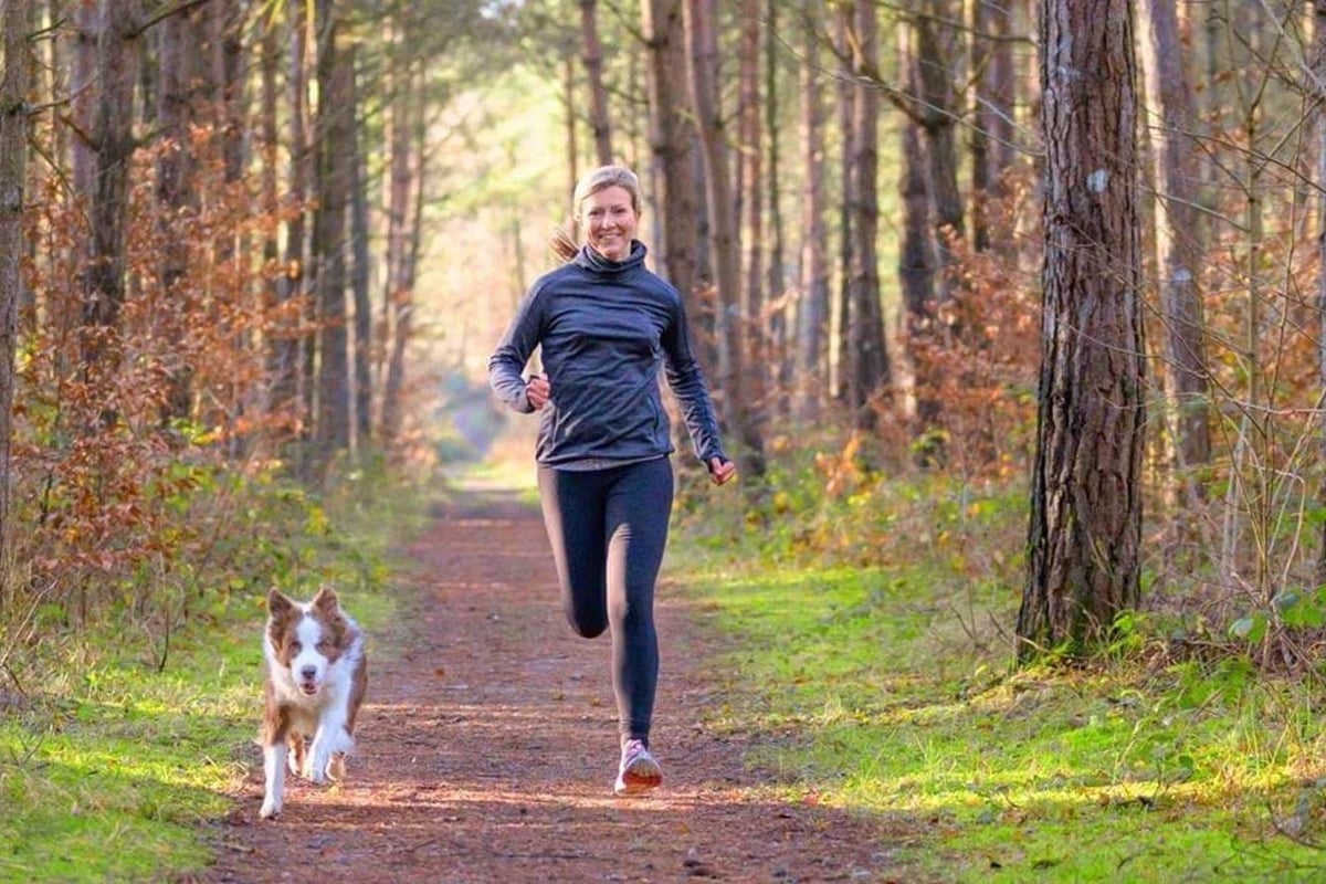 Woman jogging with dog on forest trail surrounded by tall trees and autumn leaves