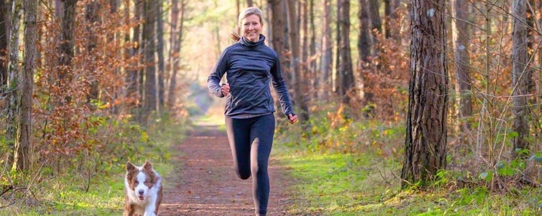 Woman jogging with dog on forest trail surrounded by tall trees and autumn leaves