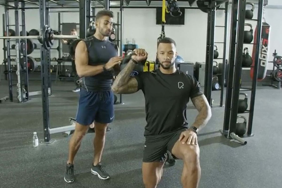 Man kneeling with kettlebell held overhead while trainer assists in gym