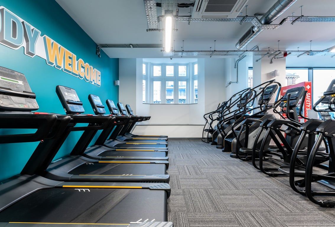PureGym London Palmers Green: Rows of treadmills and stationary cycles along teal-coloured walls in a fitness centre.