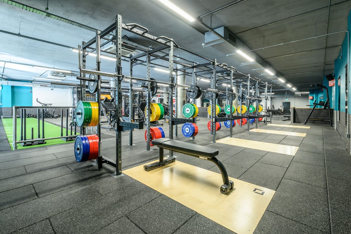 PureGym London Greenwich: Rows of squat racks, barbells, weight plates in vibrant colours, benches, and rubberized flooring.