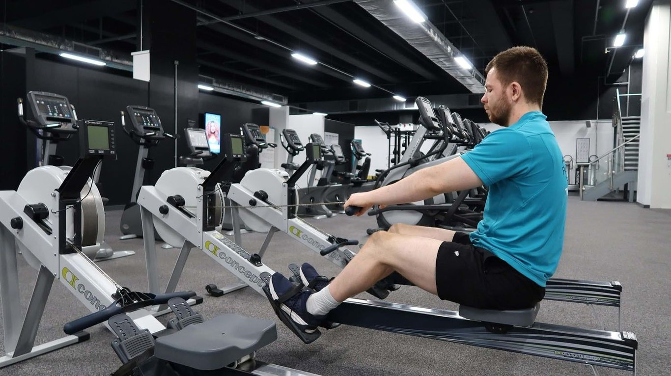 Man in blue shirt using rowing machine in gym with exercise bikes in background