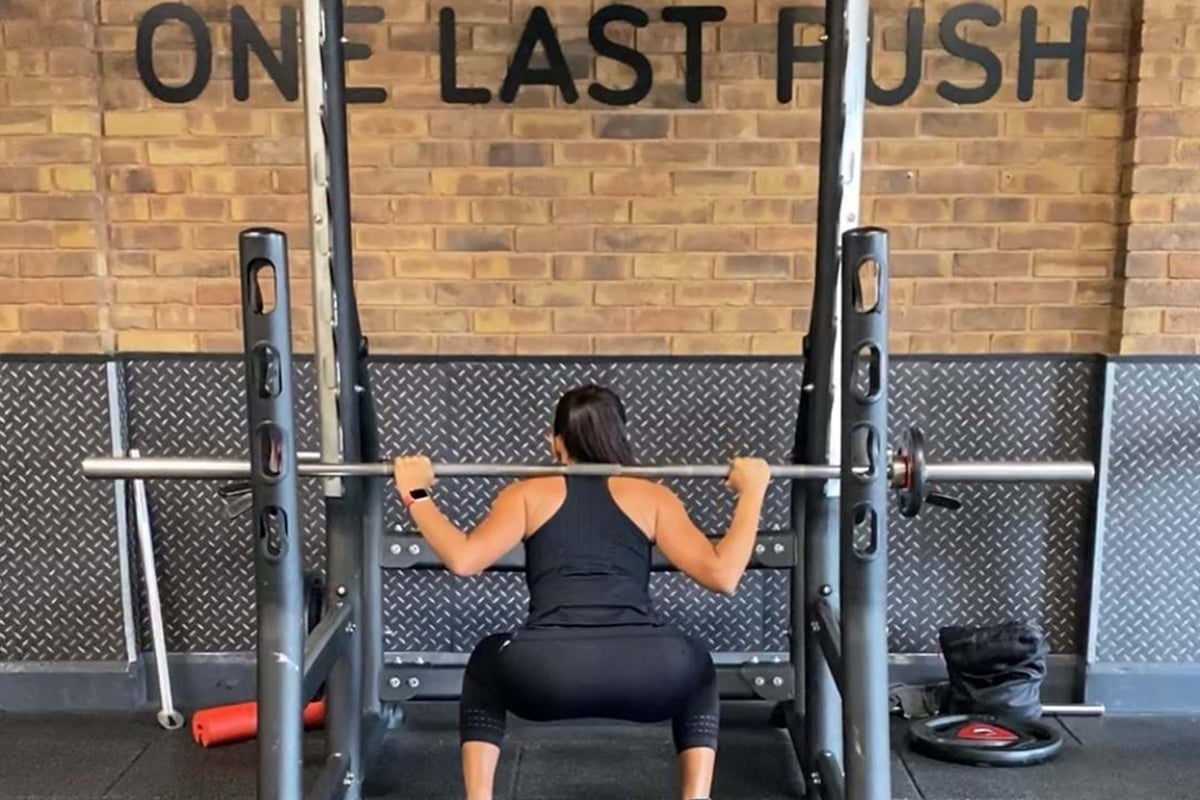 Woman squatting with barbell on shoulders inside gym rack with "One Last Push" sign behind