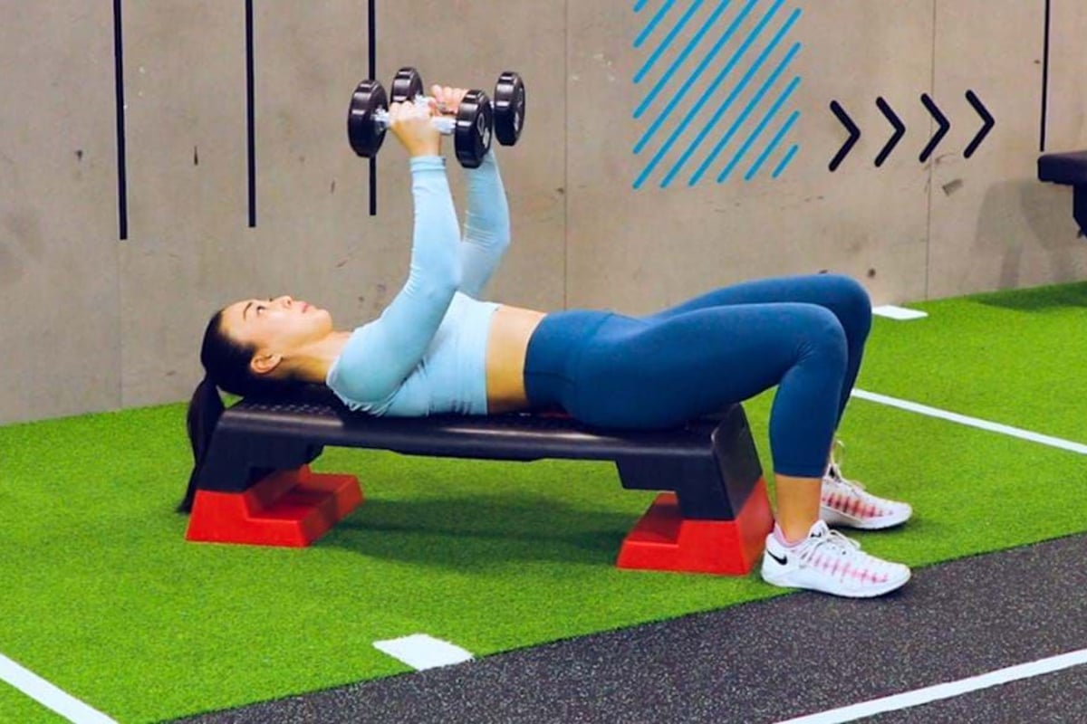 Woman in blue workout clothes lying on bench lifting dumbbells in gym