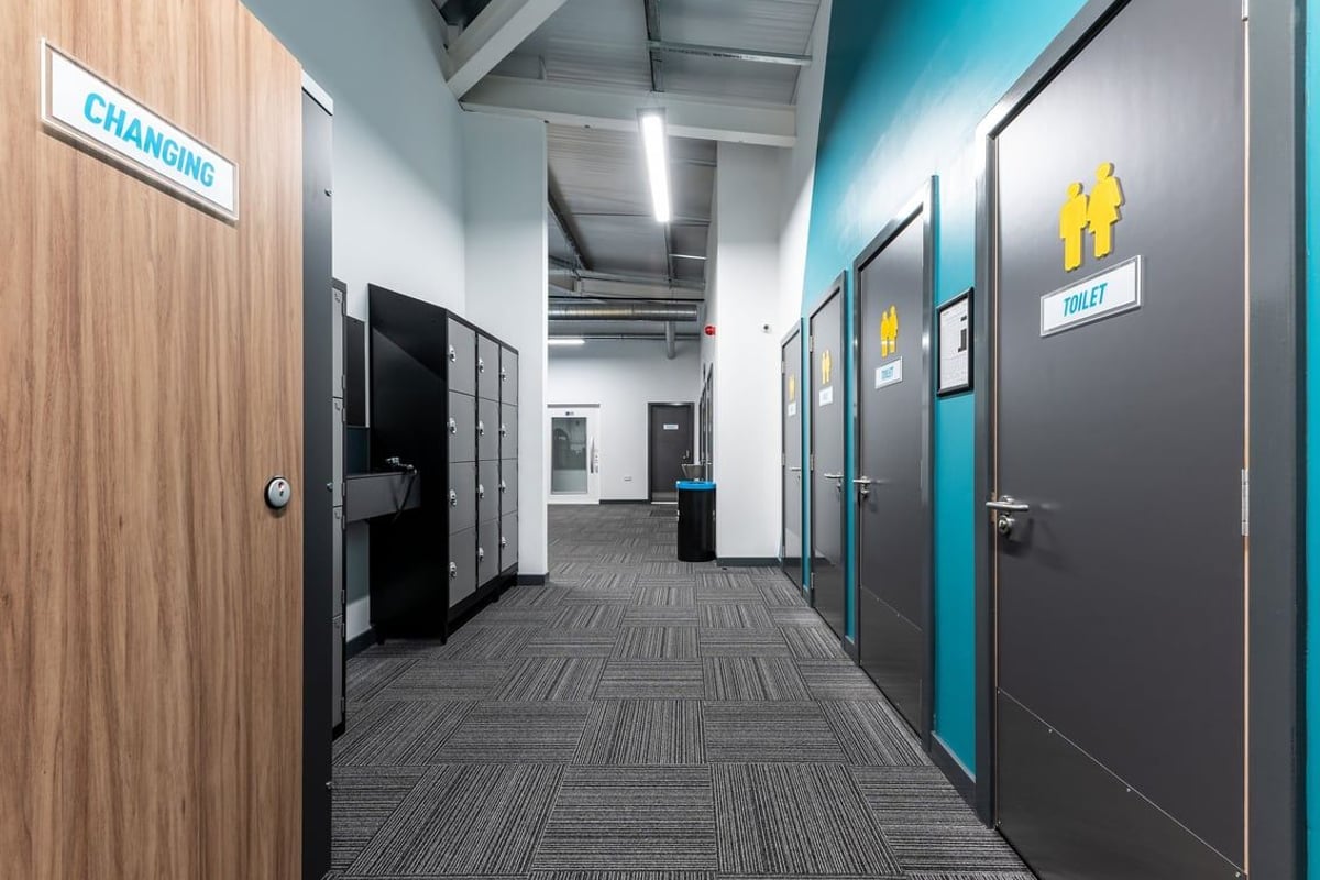 PureGym Galashiels: Changing room corridor with cubicles, carpeted floor, signage indicating facilities.