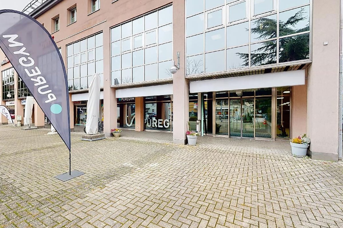 PureGym Lyss: Extérieur d'un centre de conditionnement physique avec de grandes fenêtres, un pavage en briques, des drapeaux et des bannières promotionnelles.