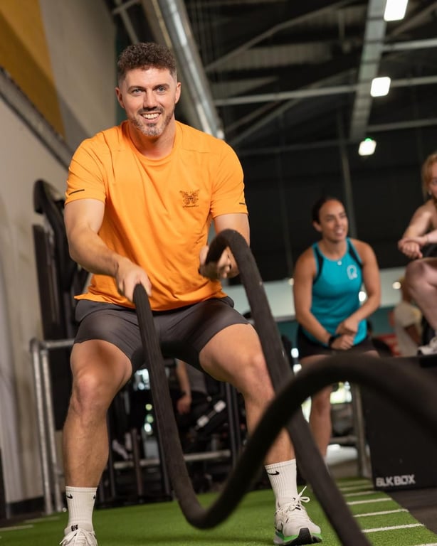 Energischer Mann im orangefarbenen Hemd, der lächelnd mit Battle Ropes in einer modernen PureGym-Anlage trainiert, während andere Mitglieder in der Nähe sind