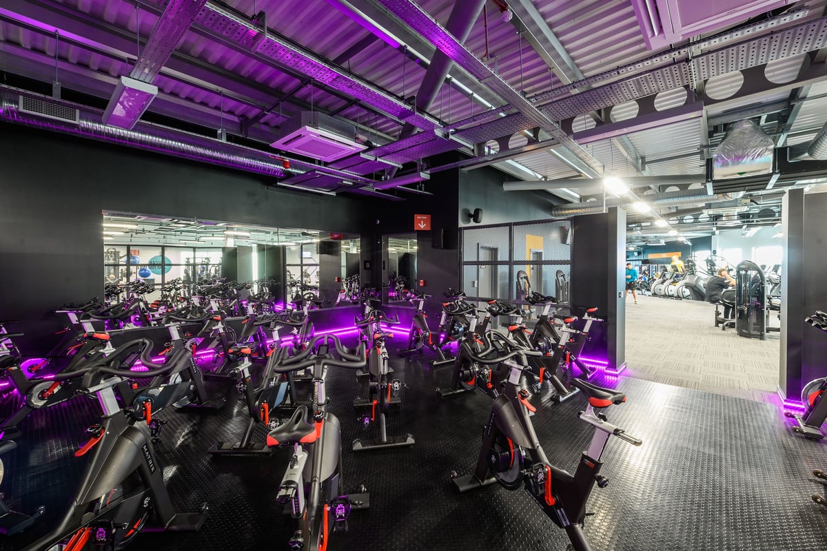 PureGym Leeds Thorpe Park: Rows of stationary bikes in a dimly lit studio with purple lighting and exposed ceiling beams.