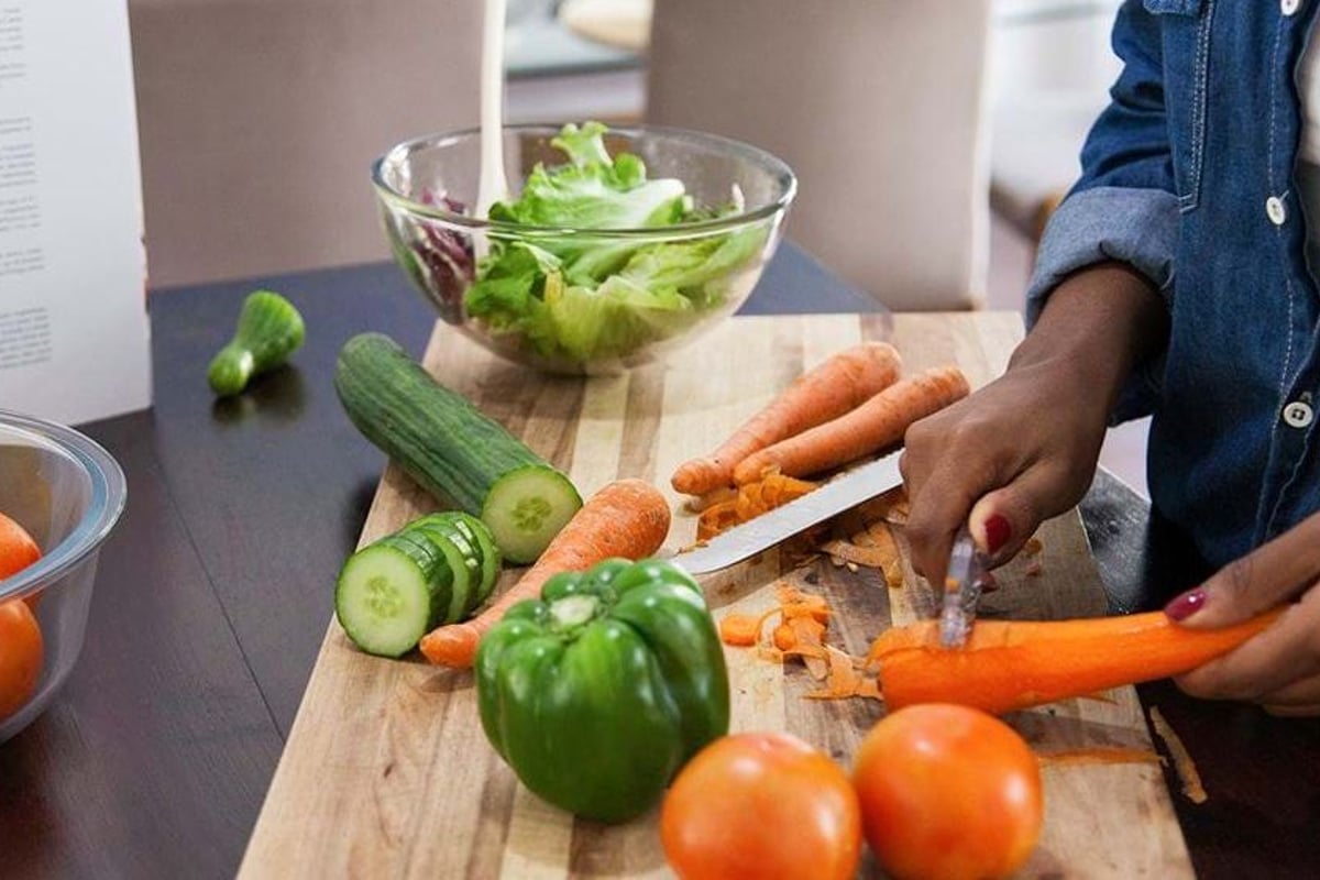 Person in denim shirt chopping carrots on a board with fresh vegetables and salad bowl