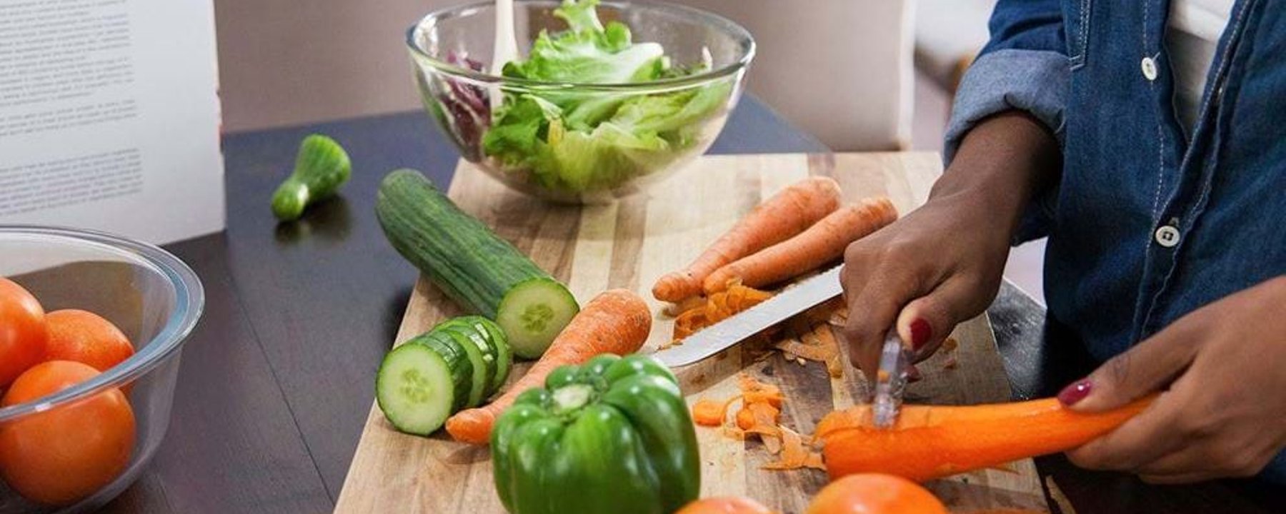 Person in denim shirt chopping carrots on a board with fresh vegetables and salad bowl