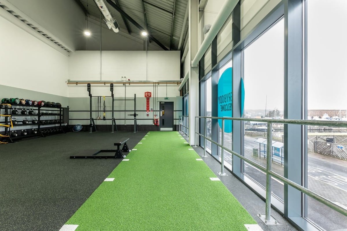 PureGym Chatham: Weightlifting area with squat racks, barbells, green turf, and mirrored wall overlooking cityscape.