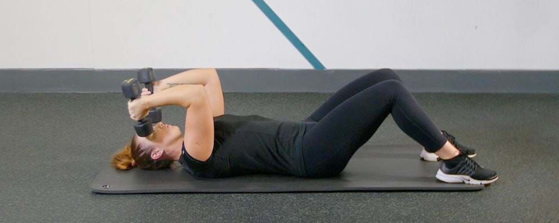 Woman lying on mat doing tricep extensions with dumbbells in gym