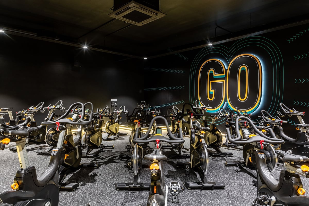 PureGym Nottingham West Bridgford: Rows of spin bikes in a dimly lit studio with neon 'GO' sign and geometric patterns.