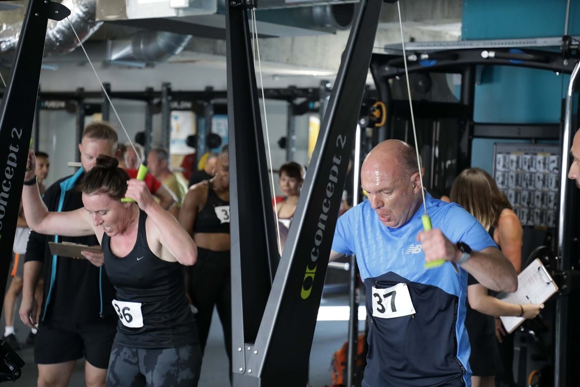 Two people intensely using Concept 2 rowing machines in a gym with others in the backgroun