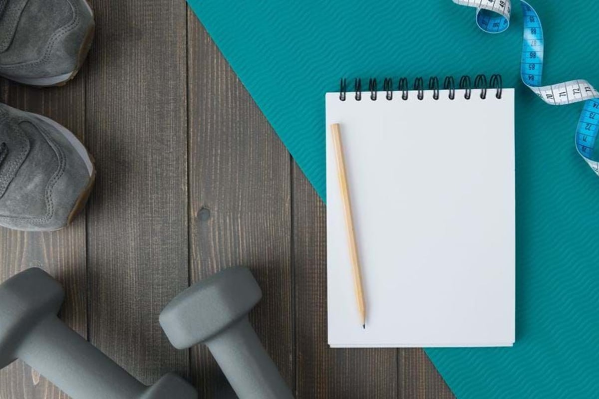 Grey trainers and dumbbells beside a blank notepad, pencil, measuring tape, and blue exerc
