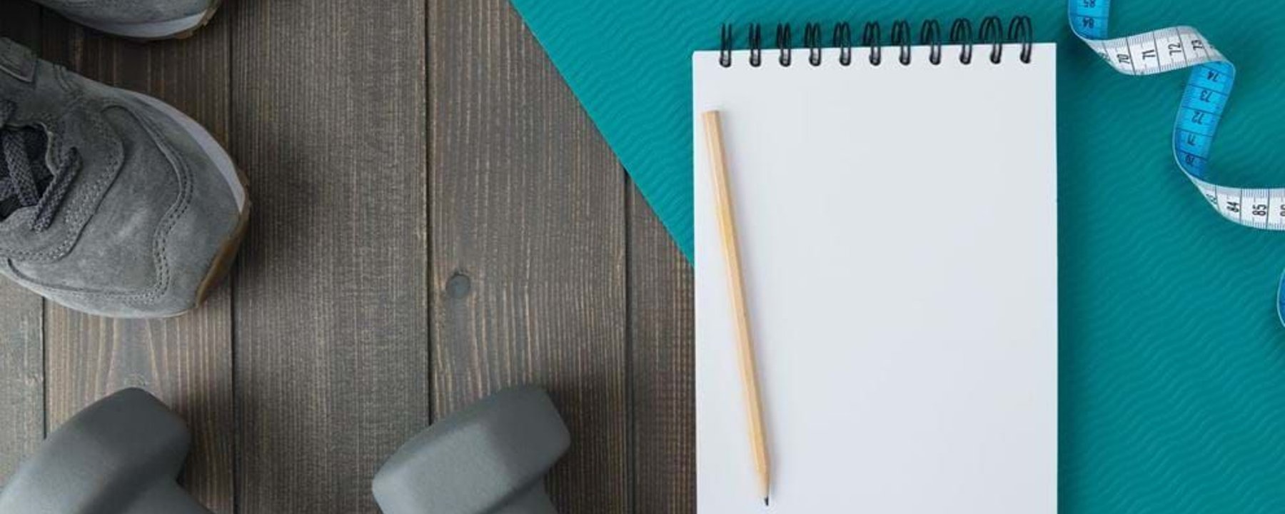 Grey trainers and dumbbells beside a blank notepad, pencil, measuring tape, and blue exerc