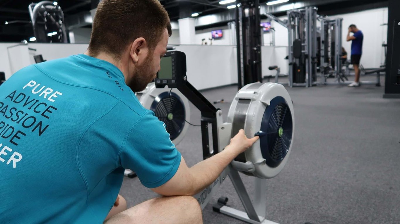Man adjusting resistance on rowing machine in gym with another person in background