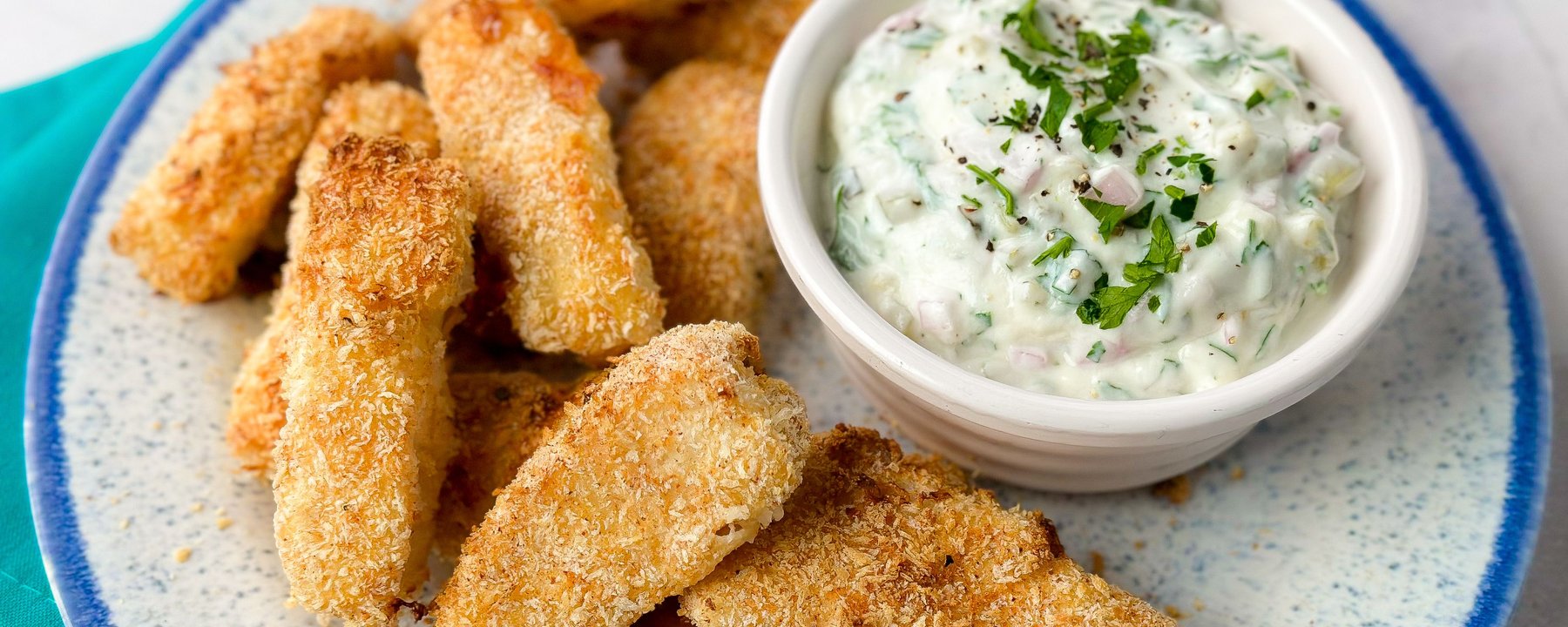 Breaded fish goujons on a plate with creamy tartare sauce garnished with herbs