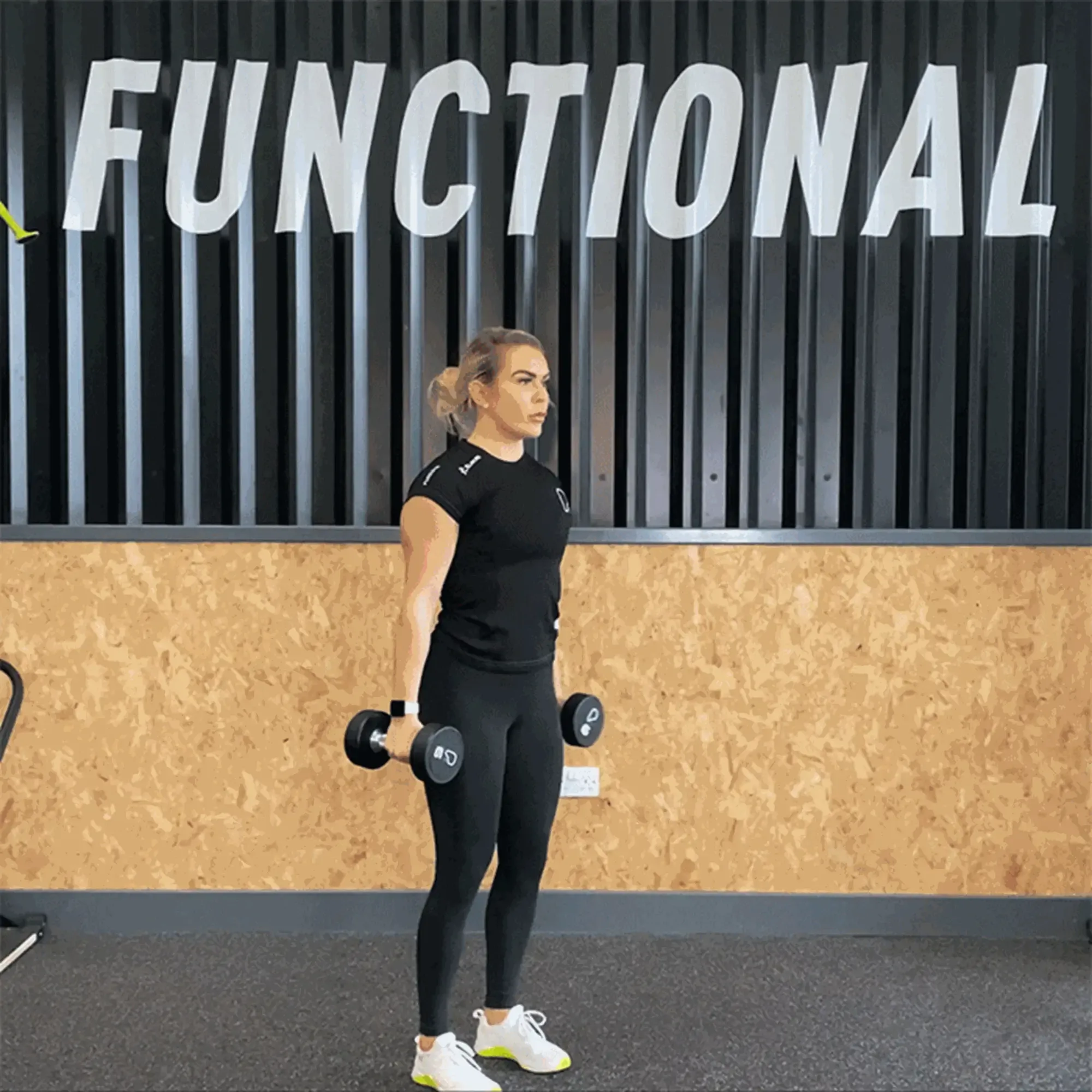 Woman in black sportswear lifting dumbbells in gym with functional sign behind her