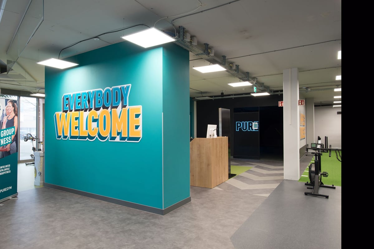 PureGym Lyss: Signalétique de bienvenue, équipement de conditionnement physique, espace ouvert avec sol caoutchouté.