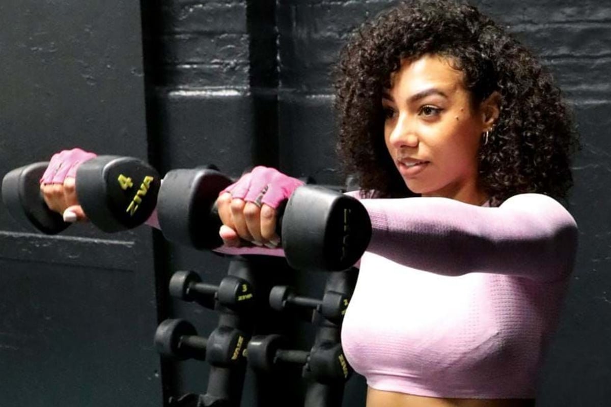 Woman in pink top lifting dumbbells in front of black wall and weights rack