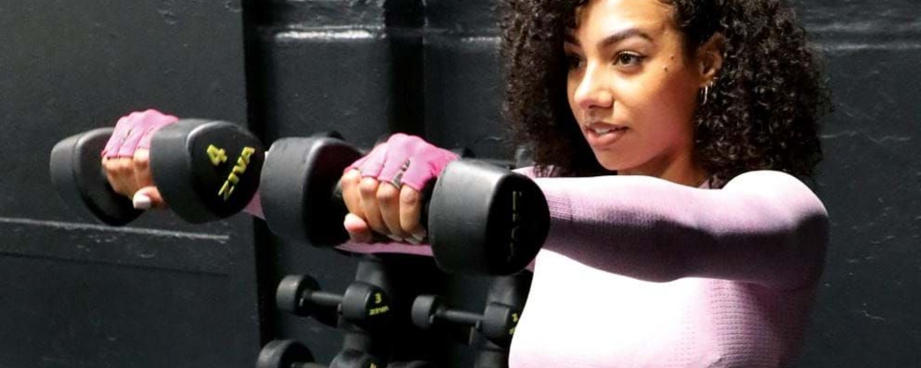 Woman in pink top lifting dumbbells in front of black wall and weights rack