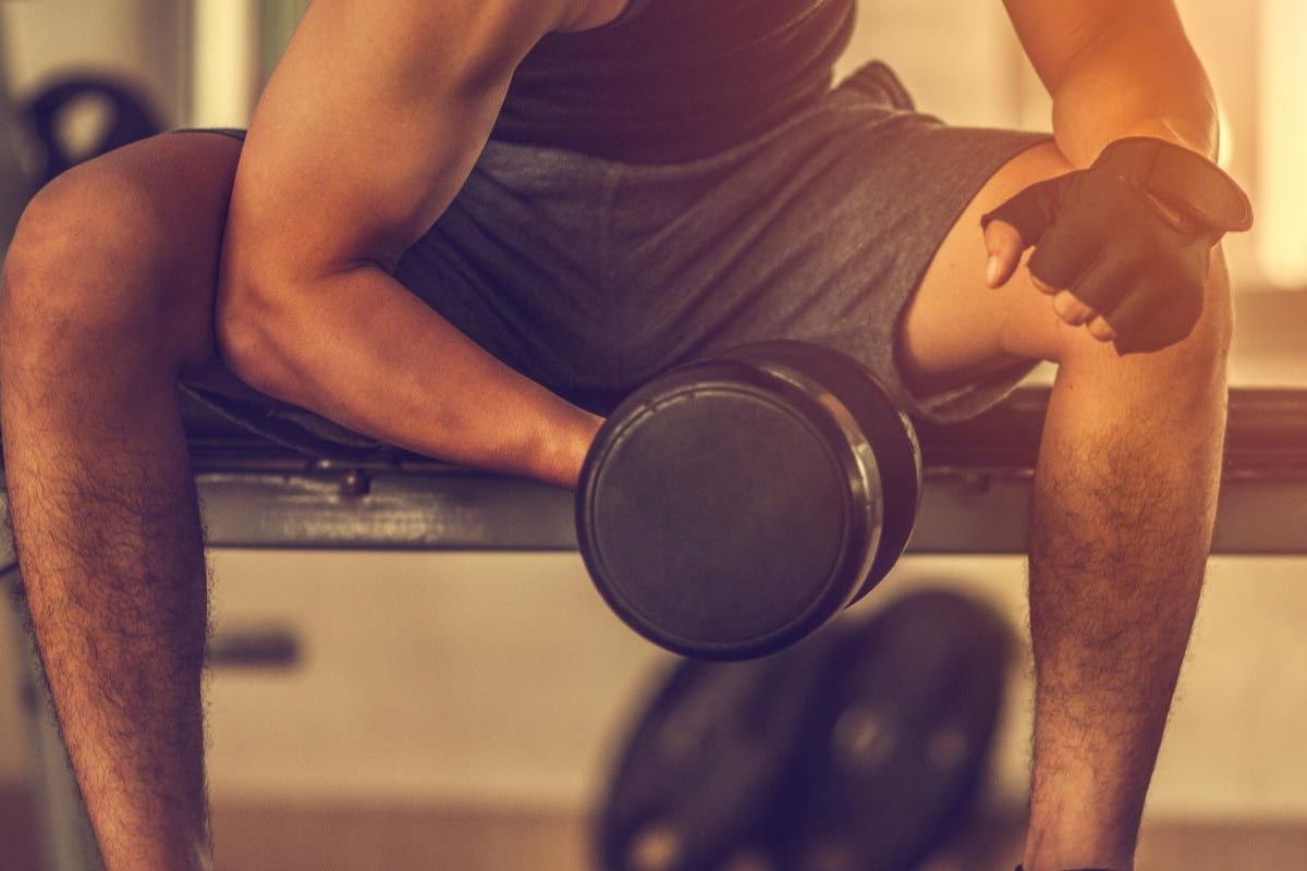 Man lifting dumbbell while sitting on gym bench wearing gloves and shorts