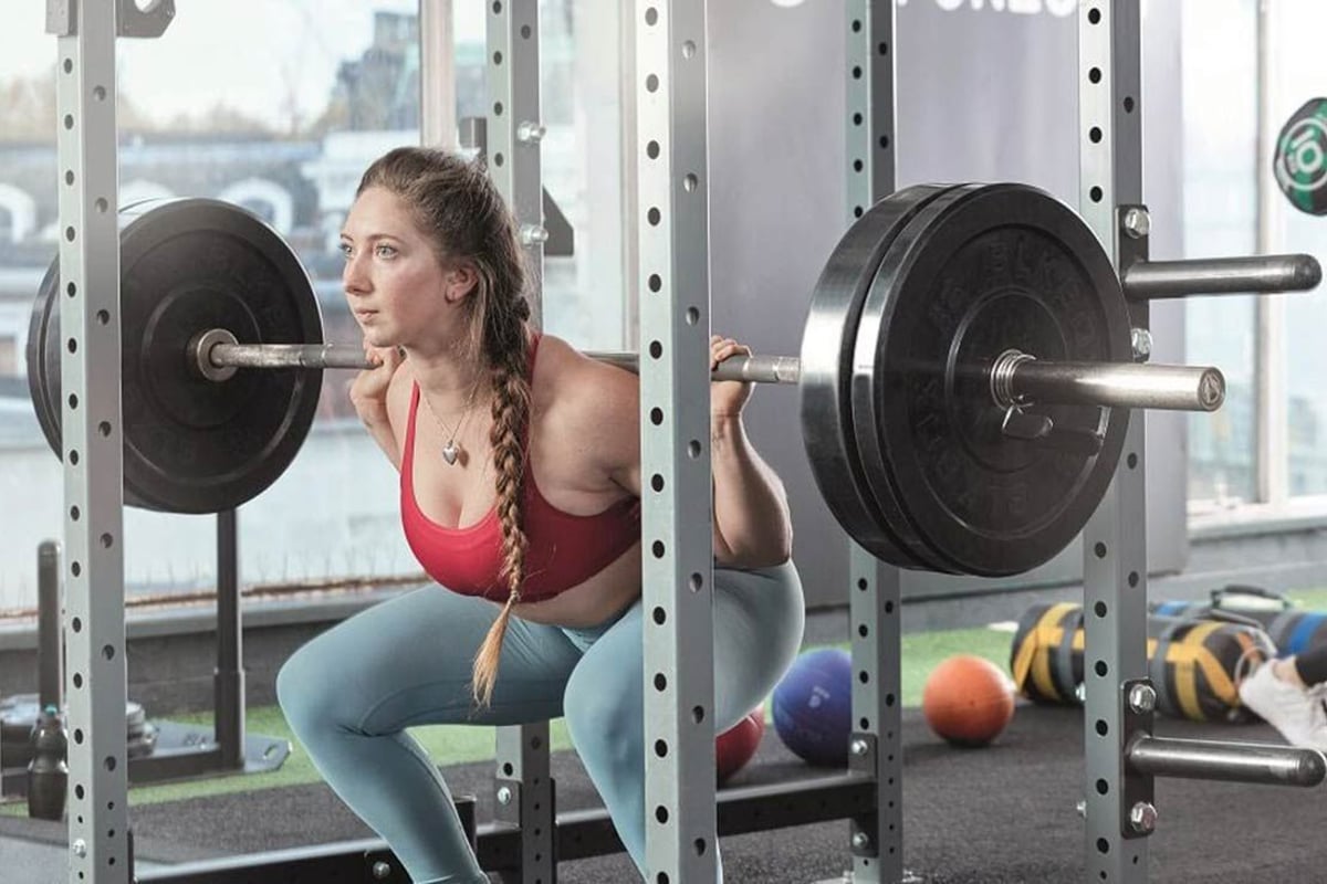 Femme en brassière rouge et legging bleu accroupie avec barre dans une cage de gym