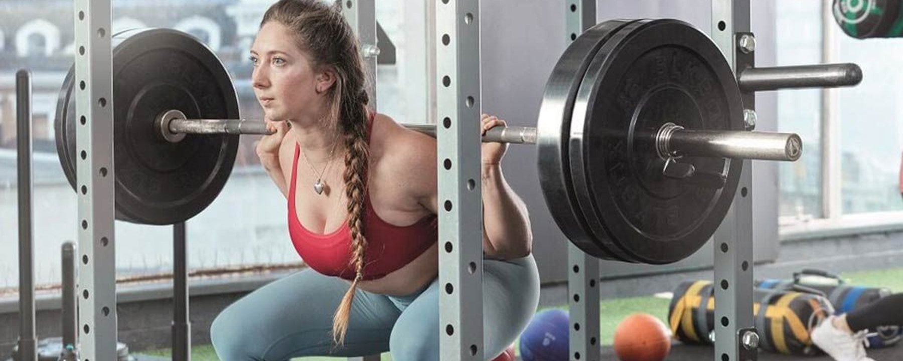 Woman in red sports bra and blue leggings squatting with barbell in gym cage
