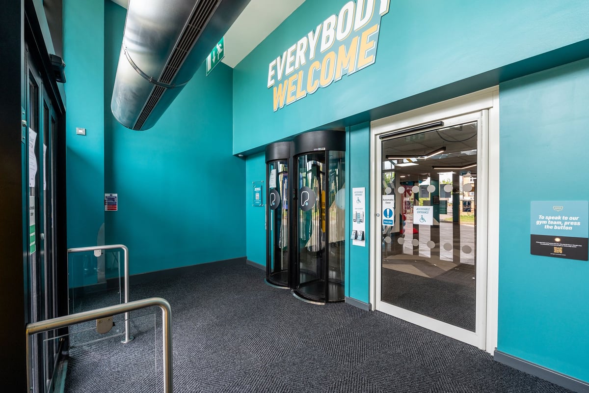 PureGym Harlow: Corridor with turnstiles, "Everybody Welcome" signage, and teal walls.