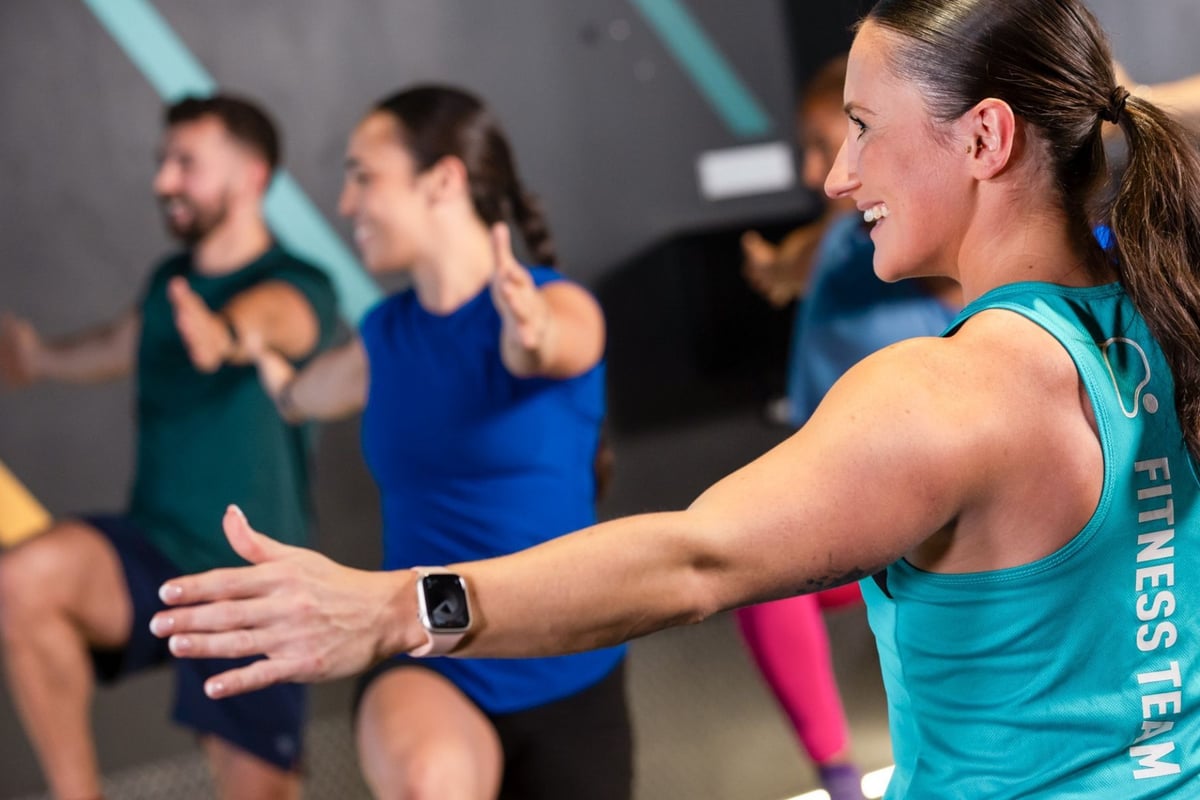 PureGym: a HIIT class lead by a female instructor stretching out her arm, with a female and a male member raising their knees in the background