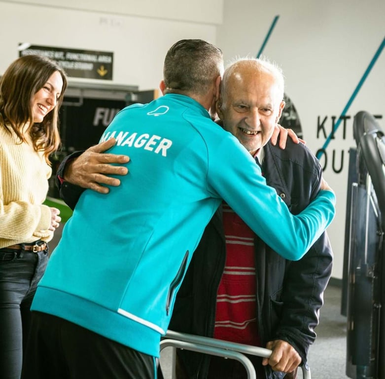 Man in blue jacket labelled manager hugging elderly man with walker while woman smiles nea